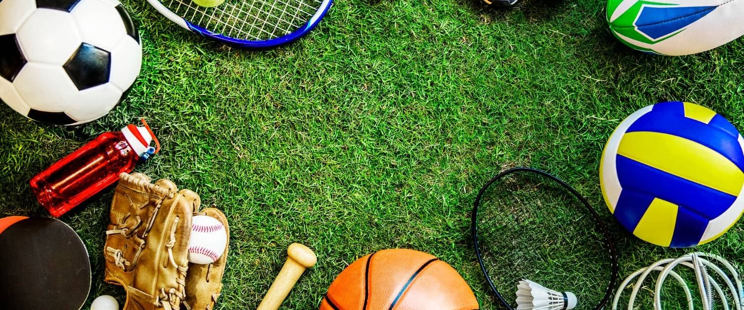 Equipment from across a school sports programme laid out on grass: soccer, tennis, baseball, basketball, volleyball, rugby, badminton and running