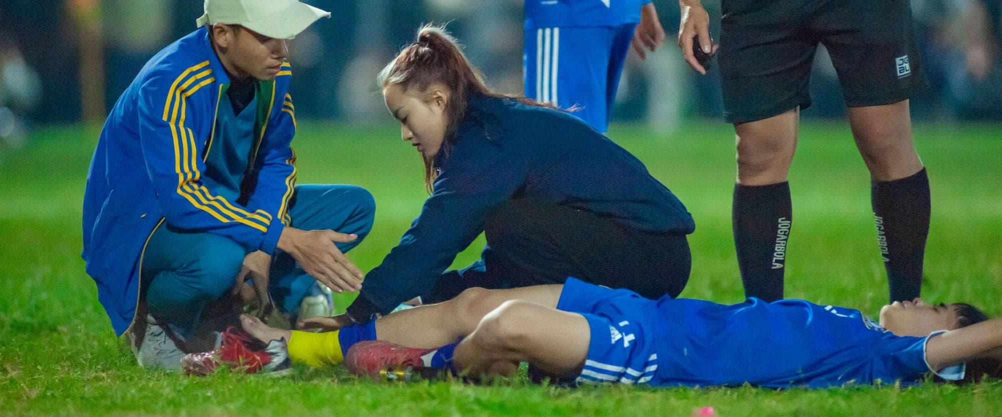 Two members of athletic staff kneeling over a player lying injured on the pitch, with the referee standing by